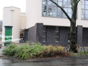 Bild: Eingangsbereich im Hintergrund. Strasse mit Mittelstreifen und Baum im Vordergrund.