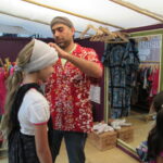 Bild: Auf dem Kleiderbasar probiert ein Mädchen ein Kopftuch, unterstützt vom Händler. Ein anders Kind steht im Vordergrund.