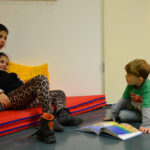 Bild: Zwei Mädchen liegen in der Kuschelecke, ein Jung kniet mit einem Buch davor.