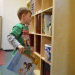 Bild: Kind steht vor Bücherregal, mit einem Buch in der rechten Hand und schaut auf andere Bücher im Regal.