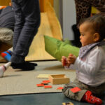 Bild: kleines dunkelhäitiges Kind spielt mit Holzkarten auf dem Fußboden. Im Hintegrund Füße und Beine von Kindern und Erwachsenen