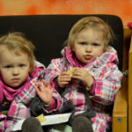 Bild: Zwilinge sitzen nebeneinander auf einem Stuhl. Kind links winkt, Kind rechts ißt Kuchen