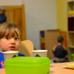 Bild: kleiner Junge sitzt bei Kaffee und Keksen am Tisch und schaut in die Kamera, im Hintergrund spielen Kinder auf dem Fußboden