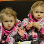 Bild: Zwillingsmädchen sitzen nebeneinander auf einem Stuhl und grüßen