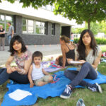 Bild: Zwei Mädchen und ein kleiner Junge sitzen auf eine Decke auf der Wiese und essen