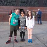 Aufgeregtes Warten, dass es endlich nach oben geht. | © FZM Bild: 3 Mädchen stehen am Fuß der Siegesäule und blicken in die Kamera