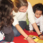 Bild: Kinder und Erzieherin beim Geschichten lesen knieend auf dem Fussboden, ein Buch in der Mitte