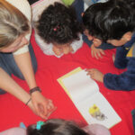 Bild: Kinder und Erzieherin beim Geschichten lesen knieend auf dem Fussboden, ein Buch in der Mitte