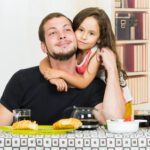 Bild: Mädchen umarmt ihren Vater von hinten am Frühstückstisch