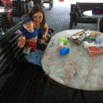 Bild: Sylvia und Benjamin machen Pause, sitzend am Tisch mit Lebensmitteln und essen. Blick zum Betrachter.