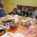 Bild: Kinder und Erwachsene sitzen um einen Tisch und basteln gemeinsam. Getränke, Obst und Kekse in der Mitte.
