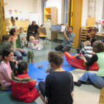 Bild: Babys, Kinder und Erwachsene sitzen auf dem Fußboden des FZM im Kreis. Mitarbeiter der Kurt-Tucholsky-Bücherei hält ein Buch mit Bildern hoch und in die Runde.