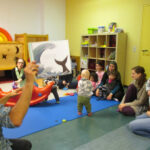 Bild: Babys, Kinder und Erwachsene sitzen auf dem Fußboden des FZM im Kreis. Mitarbeiter der Kurt-Tucholsky-Bücherei hält ein Buch mit Bildern hoch und in die Runde.