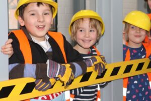 Bild: Kinder in Bauarbeiterkleidung stützen sich mit den Oberarmen auf einer Absperrung ab. Blick in Richtung Betrachter.