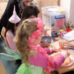 Foto: 3 Mädchen bemalten je einen Pappteller für Masken auf einem Tisch. Am hinteren Tischende junge Frau mit Kopftuch erklärt. Blick von seitlich und hinten.