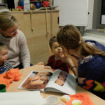 Foto: Mutter schminkt das Gesicht ihres Kindes nach davor auf dem Tisch liegender Buchvorlage. Andere Mutter links danaben am Tisch schaut mit ihrem Kind zu.