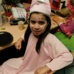 Foto: Mädchen mit blumenbemaltem Gesicht und Spitzmütze sitzt quer zwischen Tisch und Stuhllehne und blickt zum Betrachter.