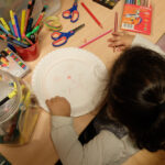 Foto: kleines Mädchen bemalt einen Pappteller für eine Maske. Blick von hinten und oben.