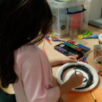 Foto: Kind bemalt einen Pappteller mit Tuschfarben für eine Maske. Blick von rechtsseitig.