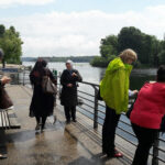 Foto: 5 Frauen am Geländer eine betonierten Seeufers. 2 schauen ins Wasser, eine auf ihr Handy und 2 unterhalten sich.