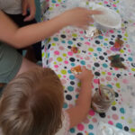 Ferienangebot - Basteln und Malen mit Gips | © FZM Foto: Kind bemalt Gipsfigur an einem Tisch. Mutter sitzt mit Baby im Arm danaben und erklärt.