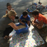 Foto: 3 Kinder und eine Erzieherin knieend und hockend auf dem Bürgersteig vor einer blauen Kiste mit Linsen, in der sich ein Schatz befindet.