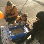 Foto: Eltern und ein Kind hockend und sitzend auf dem Bürgersteig vor einer blauen Kiste mit Linsen, in der sich ein Schatz befindet. Das Kind hat gerade einen Schatz gefunden.