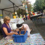 Foto: eine Erzieherin sitzend vor und ein kleines farbiges Mädchen gebeugt über einer blauen Kiste mit Linsen auf dem Bürgersteig, in der sich ein Schatz befindet. Hintergrund: der Stand vom Familienzentrum.