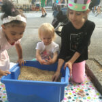 Foto: 3 kleine Mädchen knieend auf dem Bürgersteig vor einer blauen Kiste mit Linsen, in der sich ein Schatz befindet. Die Straße mit Festbesuchern im Hintergrund