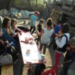 Foto: Viele Jungs und Mädchen sitzen oder stehen an einem Tisch und jeder bastelt etwas, quatscht oder liest. Bretterbude im Hintergrund.