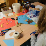 Foto: Mädchen vor einem Tisch beim Drachen basteln. Blick von hinten über die Schulter.