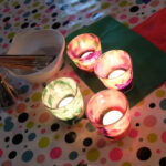 Foto: 4 Windlichter selbst beklebt mit buntem Papier und brennenden Teelichten darin auf einem Tisch (von oben). Glas und Schale mit Pinseln im Hintergrund.