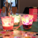 Foto: 4 Windlichter selbst bekelbt mit buntem Papier auf einem Tisch. Glas mit Pinseln und Klebstoffpackung im Hintergrund.