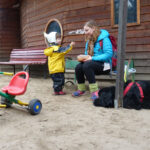 Ein kleiner Snack zwischendurch! | © FZM Foto: Kleiner Jung und ein älteres Mädchen unterhalten sich vor einer Holzhütte. Dreirad und großer schwarzer liegender Hunf im Vordergrund.