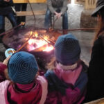 Foto: Kinder, Jugendliche und Eltern sitzen mit Stockbrotspießen vor Feuerschale. Blick von hinten über die Köpfe zweier Kinder.
