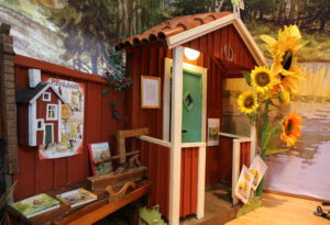 Foto: Personen-großes Model des Eingangs eines typisch schwedischen Holzhauses im rote-weißen Stil mit Sonnenblume am Vorbau. Rechts daran eine Bank mit Büchern und Werkzeug.