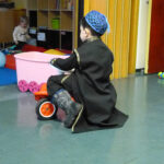 Unser Prinz vor Ort | Foto: © FZM Foto: Kind mit schwarzem Mäntelchen und Kappe auf Dreirad von hinten. Im Hintergrund spielt im Nebenraum ein anderes Kind auf dem Fussboden.