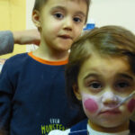 Kinder werden geschminkt | Foto: © FZM Foto: kleines Mädchen wird gerade mit Pinsel im Gesicht bemalt. Im Hintegrund schaut ein Junge zu. Beide Blick zum Betrachter.