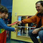 Gute Stimmung auch bei den Eltern | Foto: © FZM Foto: Kind und erwachsener Mann schäkern lachend und ziehen an der selben Tischtenniskelle. Kind im Hintergrund schaut zu.