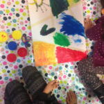 Foto: Tuschezeichnungen, Tuschefarbtöpchen und Kinderbeine auf einem Tisch mit bunt bepunkteter Tischdecke. Draufsicht von oben.