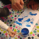 Foto: Tuschezeichnung, Tuschefarbtöpfchen blau und Kinderhand mit Pinsel darin auf einem Tisch mit bunt bepunkteter Tischdecke. Erwachsenenhand will helfen. Draufsicht von oben.