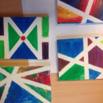 Foto: 4 Tusche-Kinderzeichnungen mit bunten eckigen Motiven auf einem Holztisch. Draufsicht von oben.