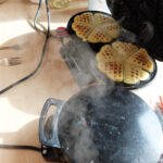 Foto: 2 Waffeleisen dampfen vor sich hin, wobei eines geöffnet ist und 2 fertige Waffelsterne zeigt.