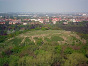 Blick auf den Drachenberg vom Teufelsberg. | © Chrischerf, Wikipedia - CC BY-SA Foto: Blick auf eine grüne abgeflachte unterhalb von Bäumen bewachsene Bergkupple. Im Hintergrund Häuser mit roten Dächern.