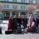 Foto: eine Gruppe Frauen unterschiedlicher Nationalität, teilweise mit Kopftuch, mit Blick zum Betrachter auf dem Bahnhofsvorplatz in Binz. Im Hintergrund rosa blühende Mandelbäume, dahinter das Bahnhofsgebäude mit dem DB-Zeichen.