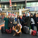 Foto: eine Gruppe Frauen unterschiedlicher Nationalität teilweise mit Kopftuch mit Blick zum Betrachter im Bahnhofsgebäude Berlin Hauptbahnhof. Im Hintergrund Fahrkartenschalter und Geschäfte.