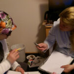 Foto: zwei Frauen, eine davon mit Kopftuch, sitzen sich gegenüber, diskutieren und gestikulieren mit einem Stif in der Hand. Auf ihrem Schoß jeweils Schreibpapiere.