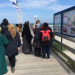 Foto: eine Gruppe Frauen, teilweise mit Kopftuch, am Stand in Rügen vor der Seebrücke mit Blick zum Wasser.
