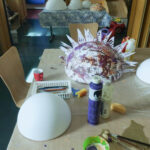 Foto: auf eine Tisch eine weiße Styropor-Halbkugel im Vordergrund, eine bemalte und beklebte Styropor-Halbkugel dahinter. Dazwischen Malzeug.