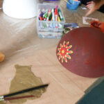 Foto: auf eine Tisch eine weiße Styropor-Halbkugel am linken Rand, dahinter und vorne je eine bemalte Styropor-Halbkugel. Dazwischen Malzeug und kretaiv werkelnde Hände.